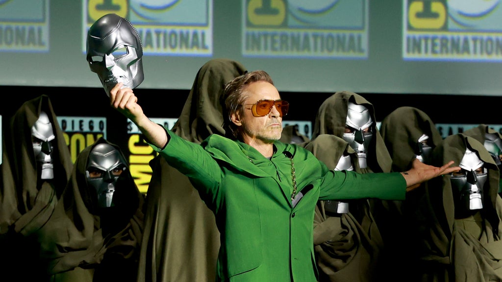 Robert Downey Jr. stands on stage at Comic-Con in a Victor Doom costume, hold his mask toward the sky in a triumphant gesture
