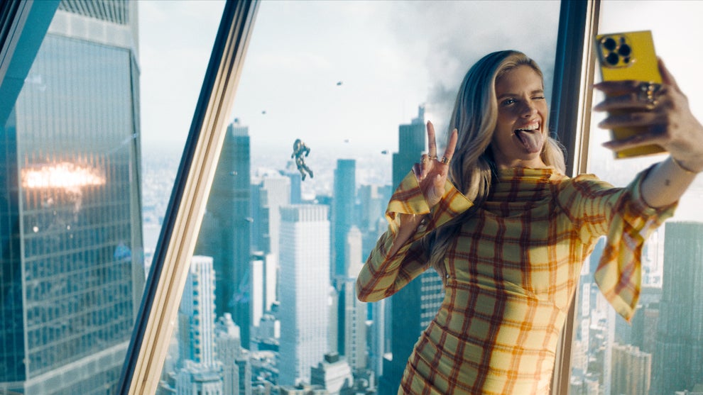 A woman stands in front of a window, taking a selfie in front of the superheroes fighting behind her