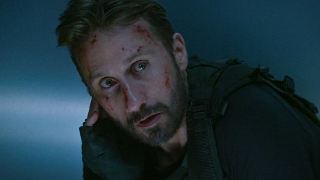 A bloodied man in tactical gear crouches in a dimly lit room, listening intently through an earpiece.