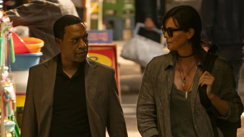 Two characters walk through a busy outdoor space, engaged in a tense conversation.