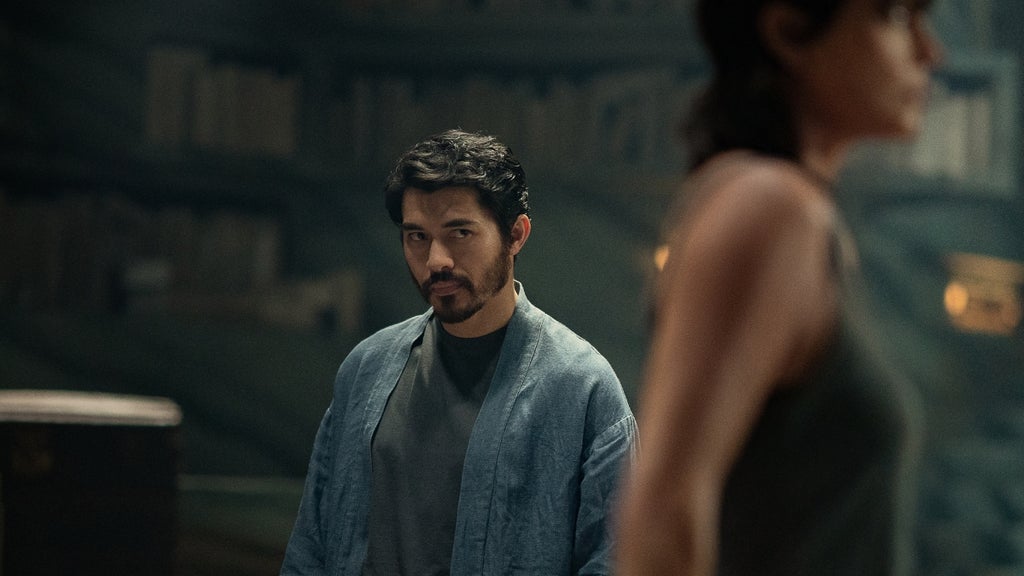 A man in a casual blue robe gazes with suspicion across the room, as a blurred female figure stands in the foreground.