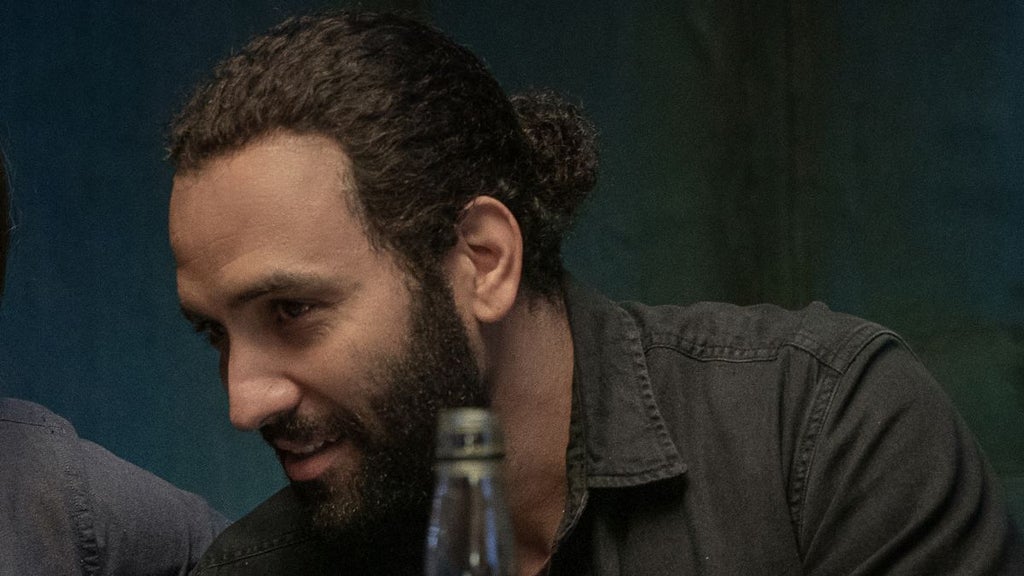 A man with a beard and pulled-back hair leans forward mid-conversation, smiling slightly