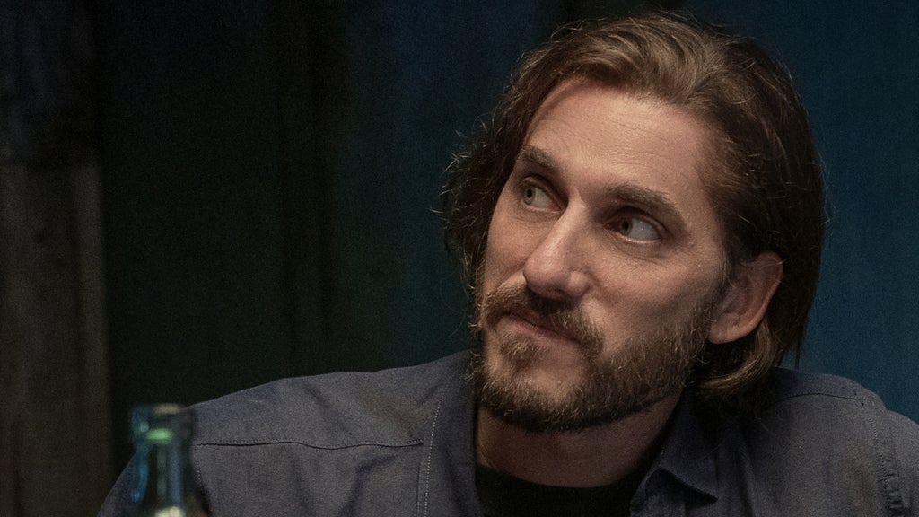 A man with shoulder-length hair and a thoughtful gaze listens attentively during an intimate gathering