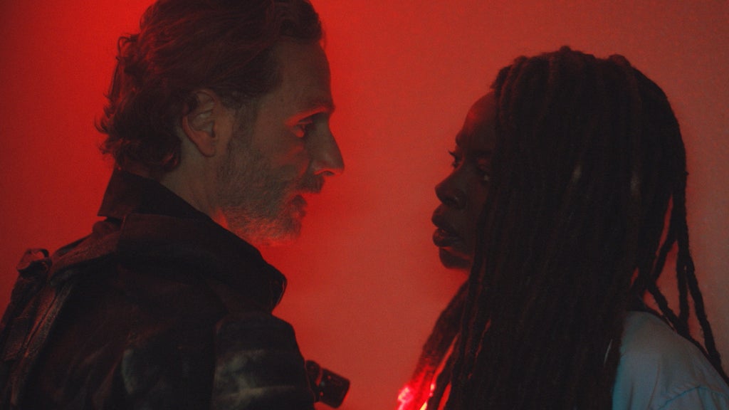 Andrew Lincoln and Danai Gurira stand close together face-to-face with dark red light flooding the frame behind them