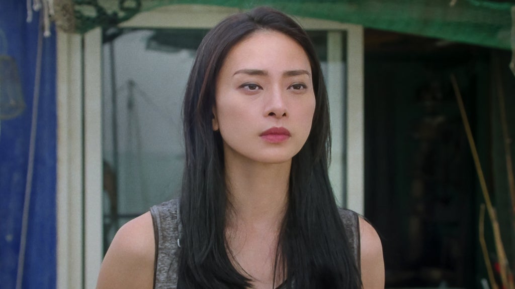 A solemn woman with long black hair stands outside a seaside structure, staring forward