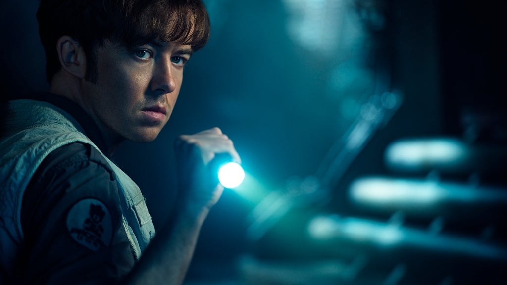 A young man in a pale jumpsuit cautiously holding a flashlight in a dim, industrial corridor.