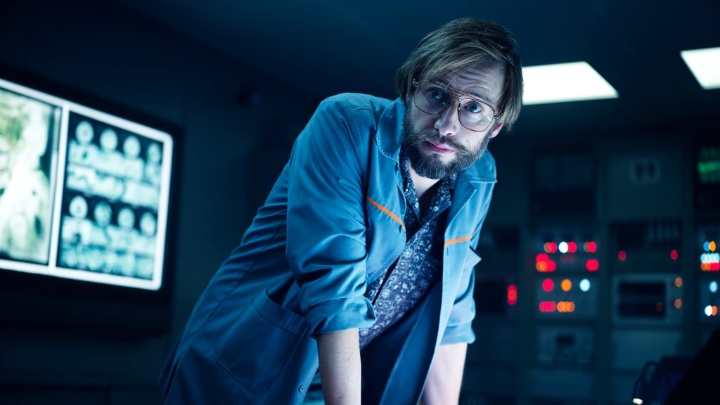 A bespectacled man in a lab coat leans over a control console, illuminated by the glow of monitors.