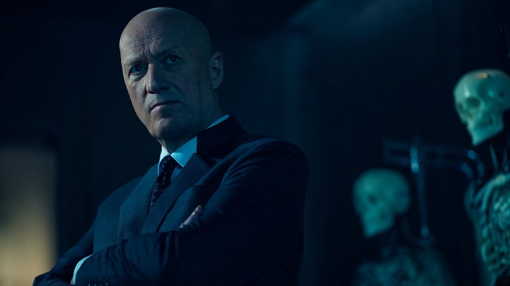 A stern, bald man in a dark suit stands with arms crossed, skeletal figures looming behind him.