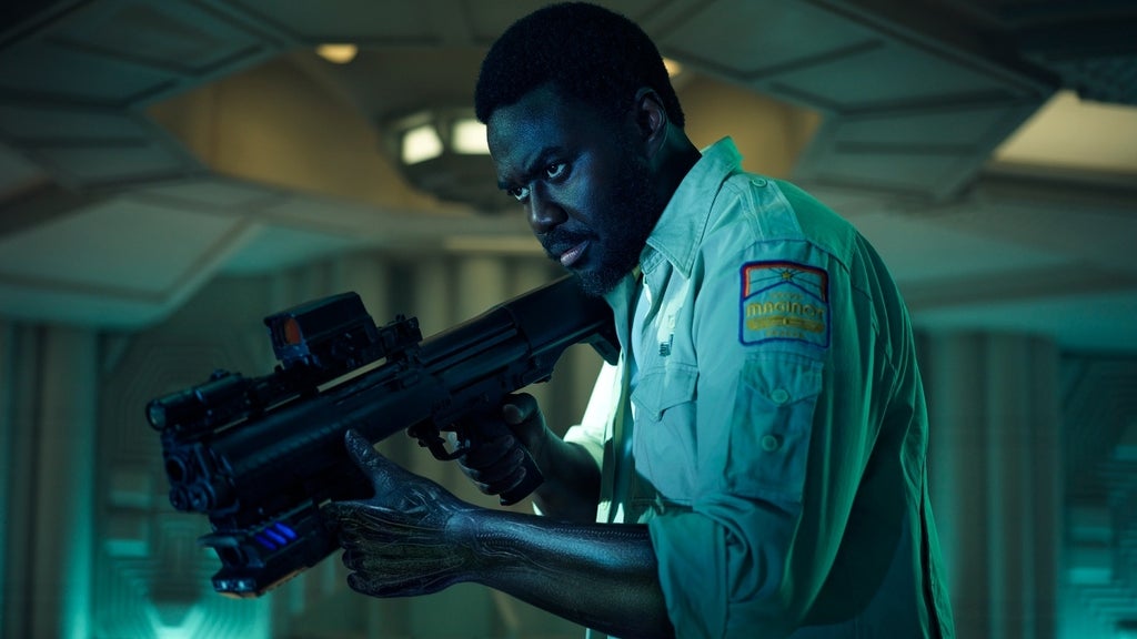 A determined man in a marine uniform aims a futuristic rifle inside a spacecraft.