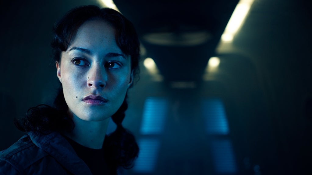 A woman with pigtails looks off to the side, standing in a dark, narrow ship corridor.