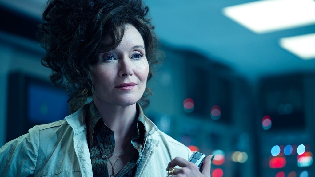 A poised woman with voluminous hair smiles faintly in a control room, holding a device.