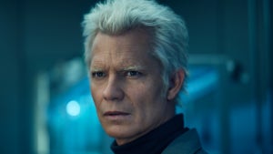 A silver-haired man with sharp features stares intently, set against a sterile, blue-lit background.