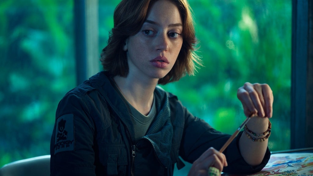 A young woman with short auburn hair sits at a table, holding a pencil, surrounded by greenery outside.