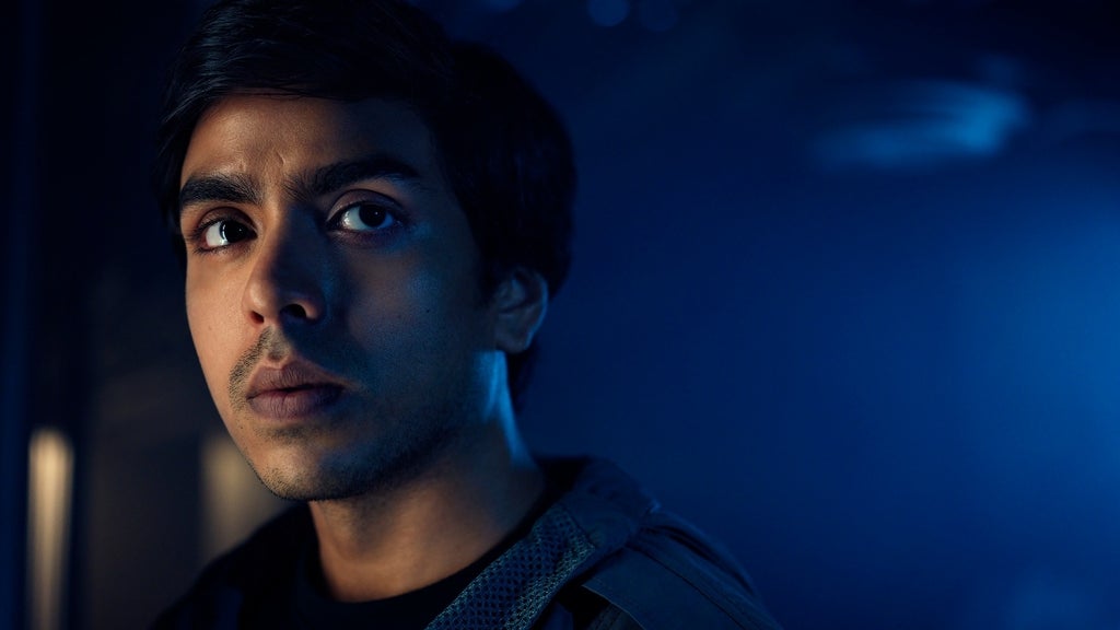 A young man in a hooded jacket looks off to the side with a wary expression, bathed in blue light.