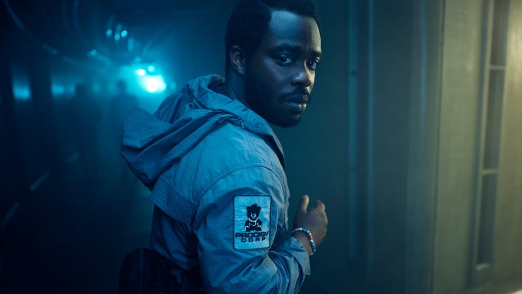 A man in a Prodigy Corp jacket glances back over his shoulder while walking down a dim, misty corridor.