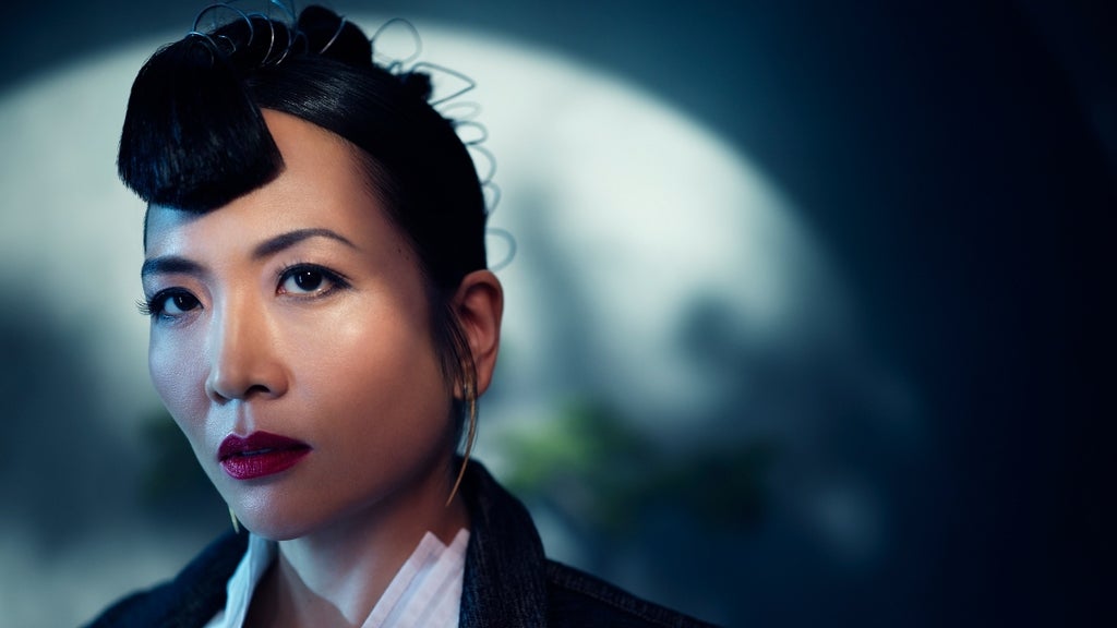 A woman with sleek, styled hair and red lipstick gazes forward under dramatic lighting.