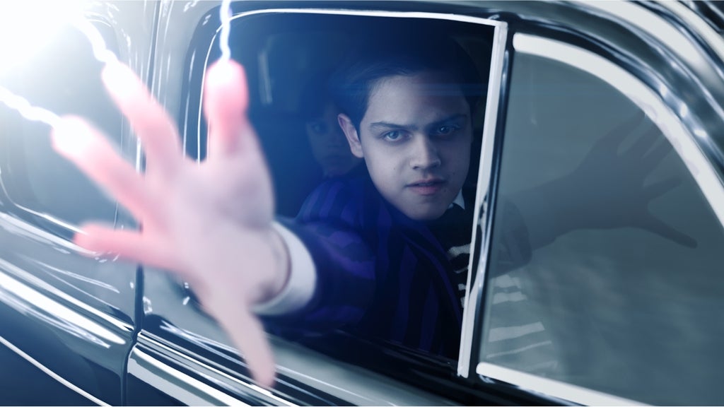 Isaac Ordonez as Pugsley Addams reaches out of a car window using his electric powers with Wednesday Addams looking on in the seat behind him