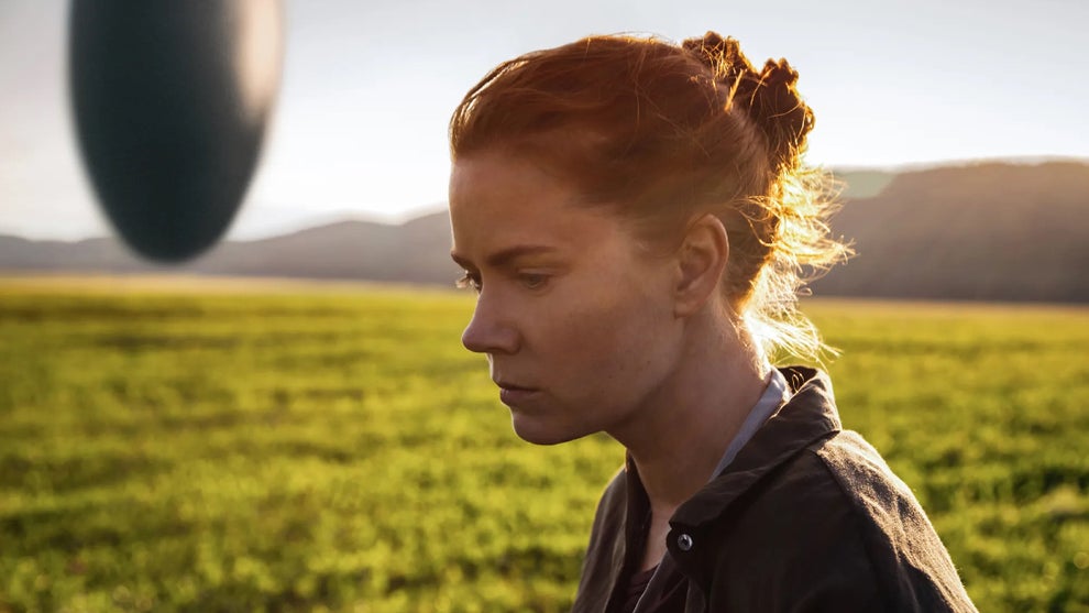 Amy Adams stands in a grassy field with an alien spacecraft in the background and the sun to her back