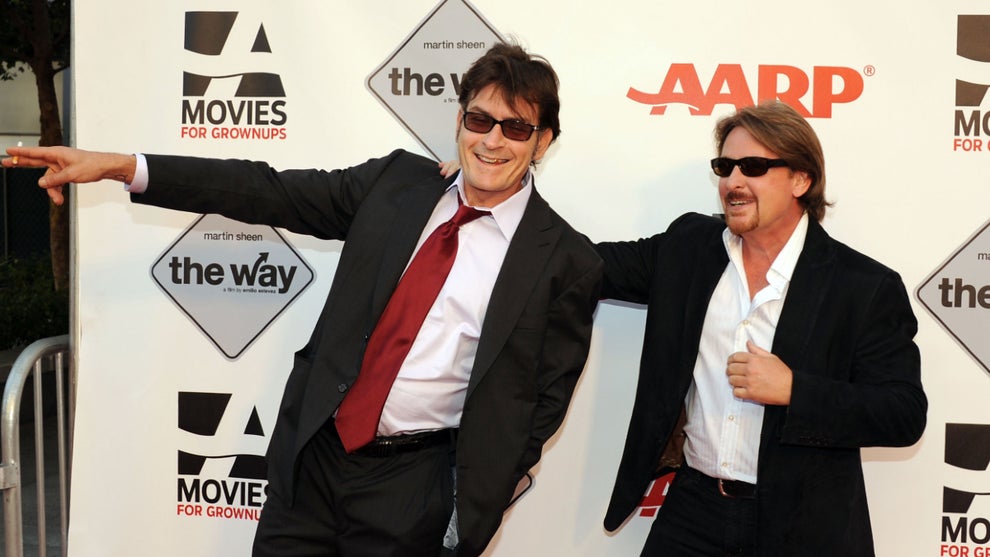 Charlie Sheen (left) and Emilio Estevez (right) wear suits and sunglasses before an AARP background. Sheen points to his right