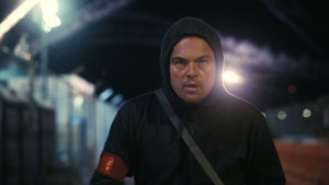 Leonardo DiCaprio walks at night in a black hoodie with fireworks behind him in "One Battle After Another"