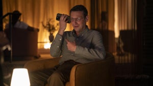 Tim Robinson as Ron sitting in a chair and looking weird while pointing at his phone that he's holding up to his year in a still from the HBO series "The Chair Company."