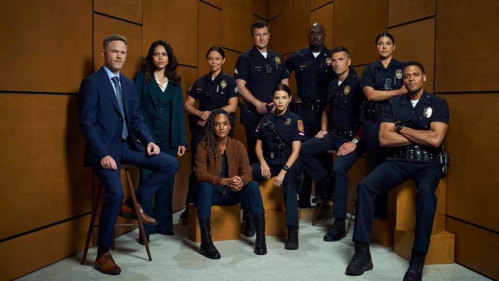 The cast of the Rookie poses for a group photo in a room with grey carpet and wood-paneled walls