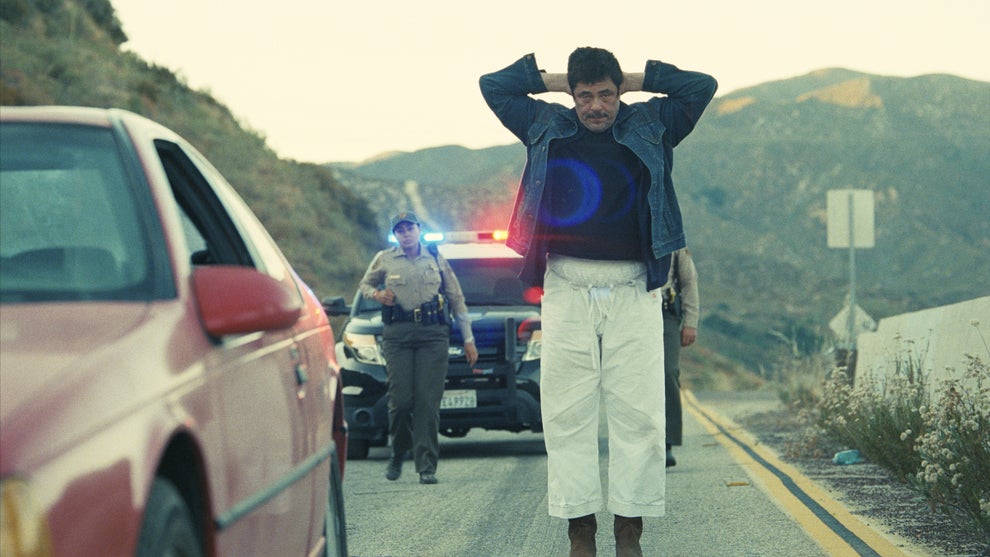 Benicio Del Toro holds his hands behind his head as he's approached by police officers in "One Battle After Another"