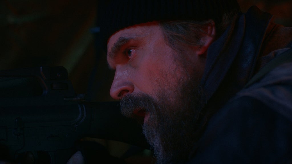 An extreme close-up of a man with a beard looking through the scope of a gun