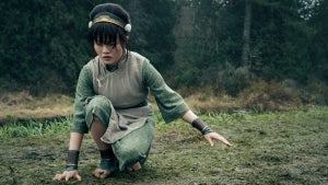 Miya Cech as Toph in "Avatar: The Last Airbender" (dressed in Toph's green clothing and gold/green headband from the animated series) crouches with one hand to the grass and the other hand held out in a wooded clearing