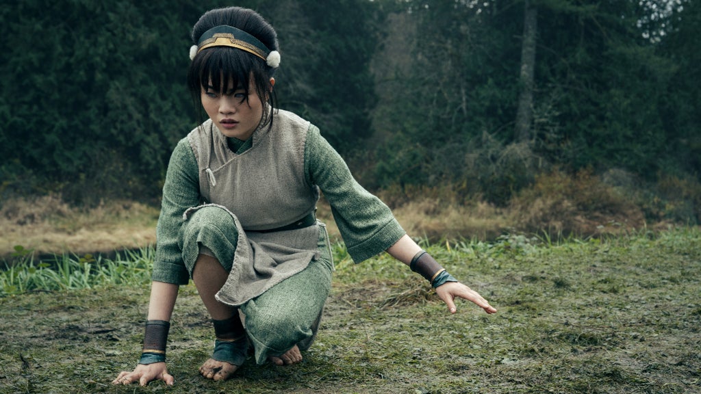 Miya Cech as Toph in "Avatar: The Last Airbender" (dressed in Toph's green clothing and gold/green headband from the animated series) crouches with one hand to the grass and the other hand held out in a wooded clearing