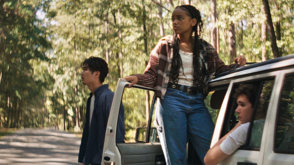 Three young adults sit on/stand near a car surrounded by forest