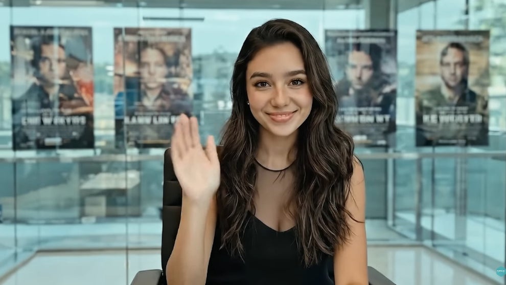 AI "Actress" Tilly Norwood waves at camera in glass room with posters behind her