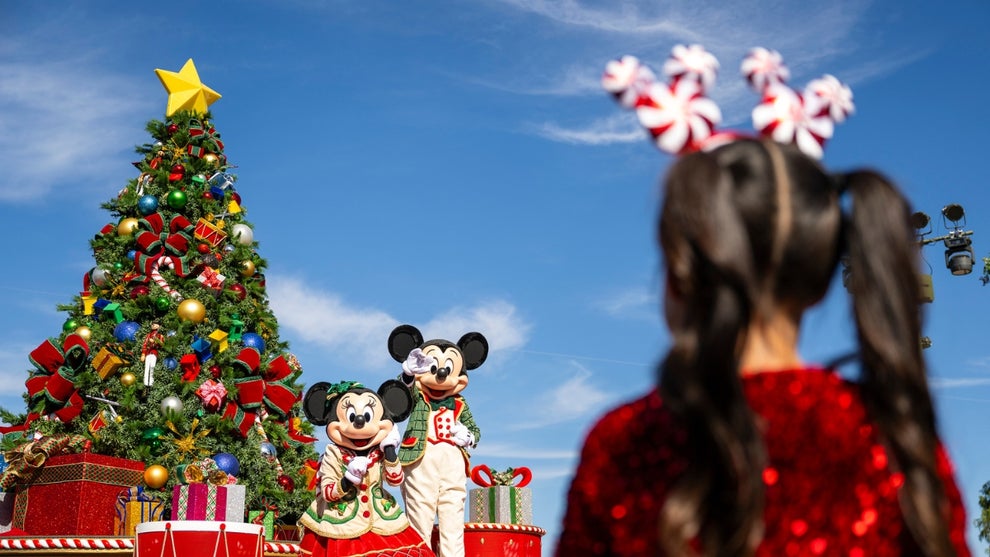 Mickey and Minnie Mouse stand in front of a Christmas Tree at the Disney Parks Christmas Day Parade while a girl with pigtails and candy-cane striped Mickey Ears looks on