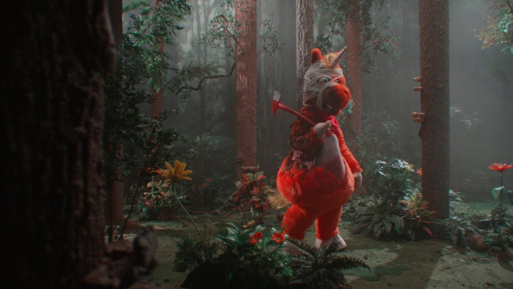 An orange uniform mascot costume stands bandaged and bloody in the woods with an ax