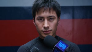 Hudson Williams looks directly into camera talking into a news microphone in "Heated Rivalry"