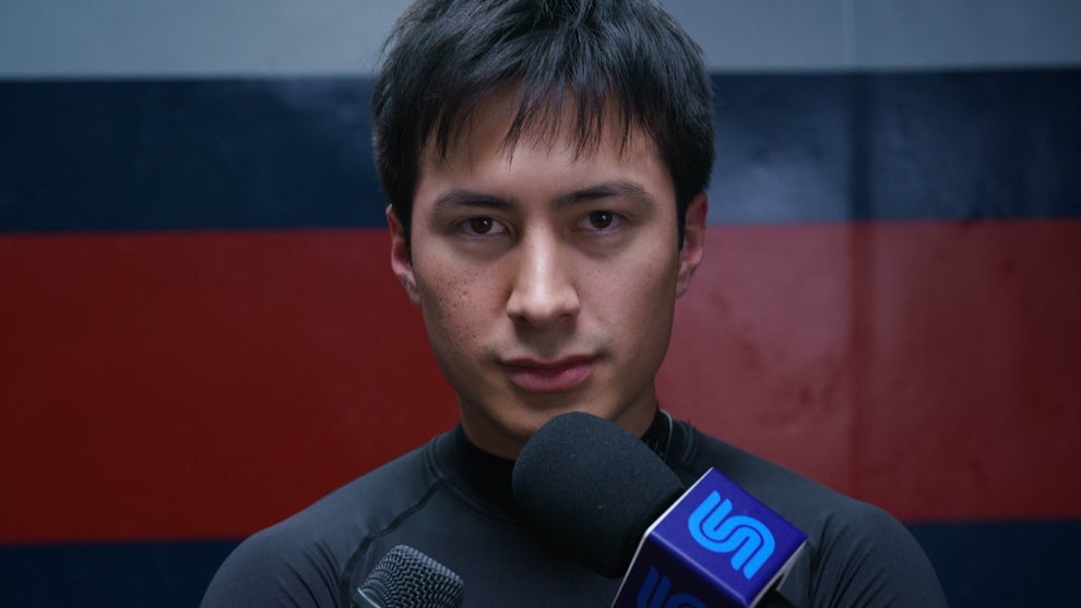 Hudson Williams looks directly into camera talking into a news microphone in "Heated Rivalry"