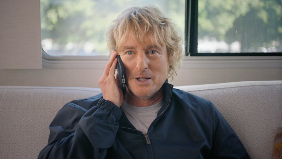 Owen Wilson sits on a couch with a phone to his ear
