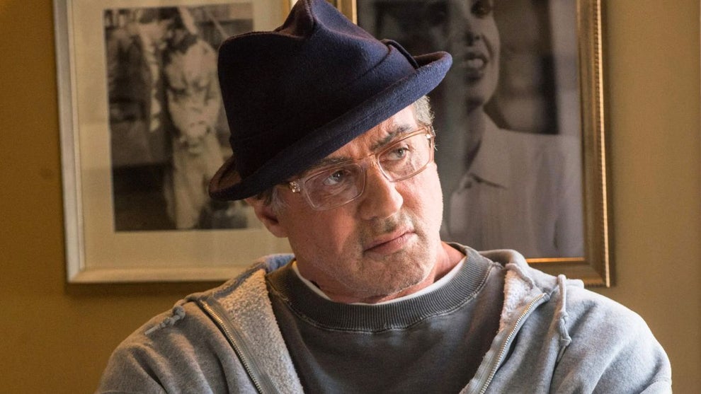 An older man wearing glasses, a dark fedora, and a gray hoodie sits at a table indoors, looking off to the side with a thoughtful expression; a glass pitcher and framed black-and-white photos are hanging on the wall in the background.