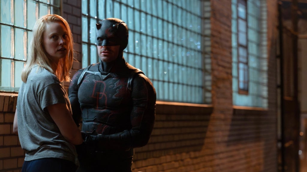 A masked vigilante in a dark suit stands beside a blonde woman outside a brick building with glowing glass-block windows at night, both looking alert and tense.