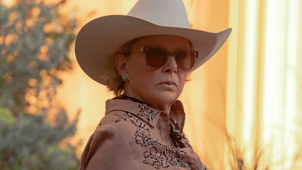An older woman wearing a white cowboy hat, sunglasses, and an embroidered western shirt looks off-camera in warm golden light outdoors.
