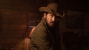 A rugged man in a cowboy hat and dark jacket stands in a dimly lit rustic cabin, looking intensely toward the camera under warm amber lighting.