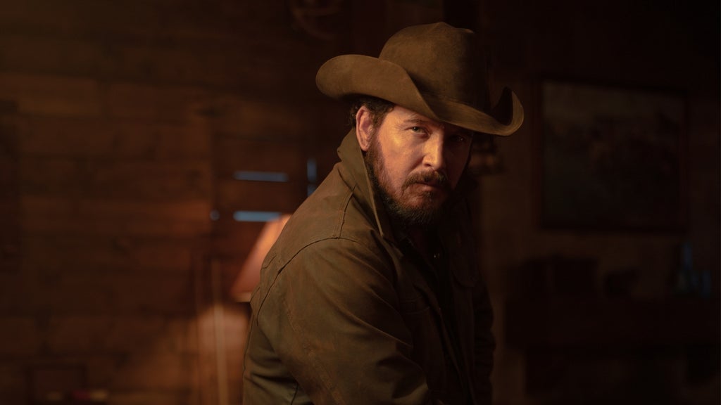 A rugged man in a cowboy hat and dark jacket stands in a dimly lit rustic cabin, looking intensely toward the camera under warm amber lighting.