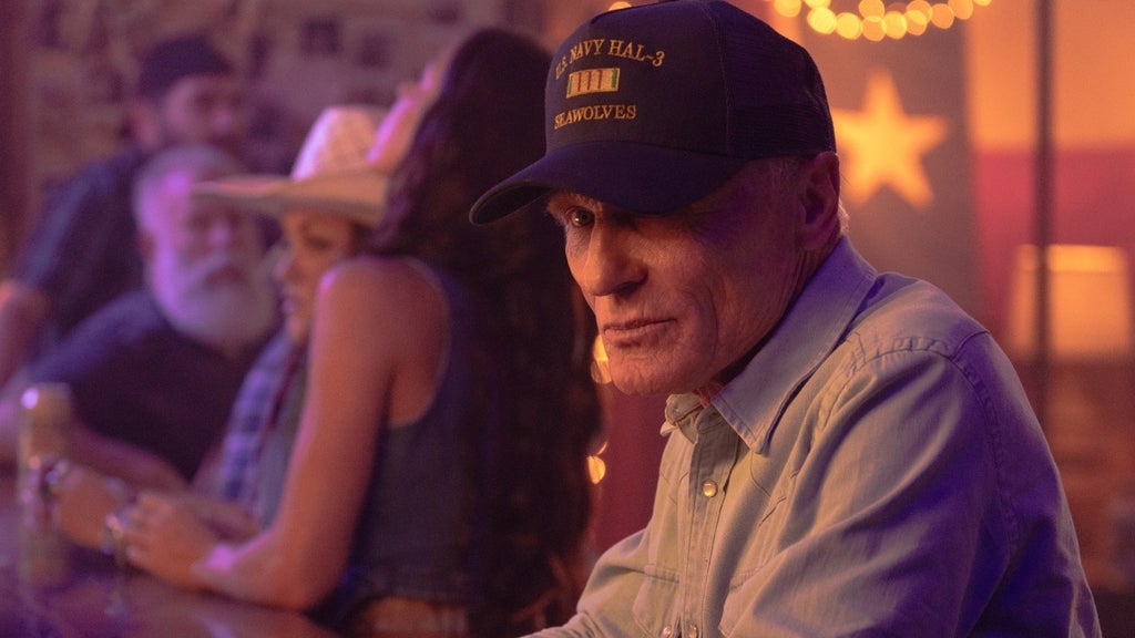 An older man sits at a bar or table in a warmly lit honky-tonk, wearing a navy blue cap reading "U.S. Navy HAL-3 Seawolves." His expression is quiet and contemplative, shoulders slightly hunched, as activity blurs in the background behind him.