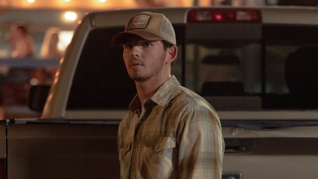  young man in a tan plaid flannel shirt and a trucker cap reading "Dutton Ranch" stands near the bed of a pickup truck at night, looking back over his shoulder with a wary expression. Warm bar or parking lot lights glow behind him. 