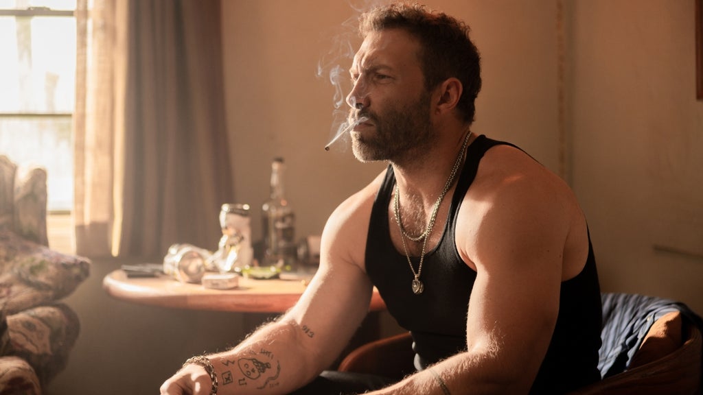 A muscular man in a black tank top sits in a dimly lit room bathed in warm, dusty light from a nearby window. He smokes a cigarette, gaze fixed in the middle distance with a brooding expression. He wears layered silver chain necklaces, a bracelet, and has visible tattoos on his forearm. A cluttered table with bottles and an ashtray sits behind him.