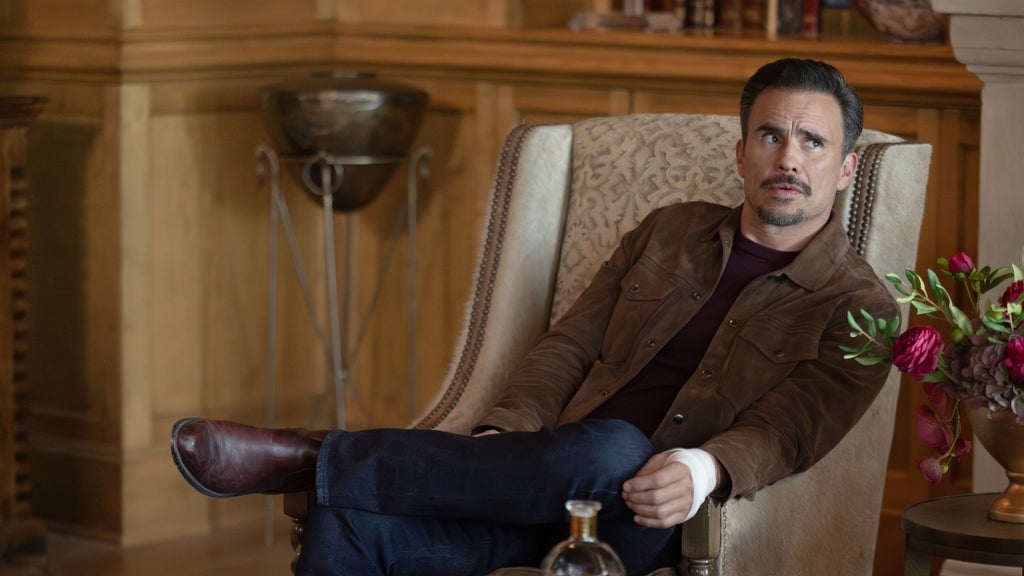 A dark-haired man with a goatee sits reclined in a cream wingback armchair, legs crossed, holding a whiskey decanter near his knee. He wears a brown suede jacket over a dark shirt and dress boots, glancing upward with a slightly guarded expression. A glass of whiskey and a vase of pink flowers sit nearby.