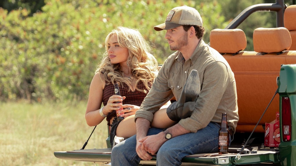 A man and woman sit side by side on the open tailgate of a vintage green Ford Bronco in a dry, grassy field. The woman, with long blonde hair, holds a drink, while the man wears a tan baseball cap and a gray button-down shirt. Beer bottles rest beside them. The scene evokes a relaxed, rural Texas setting. 