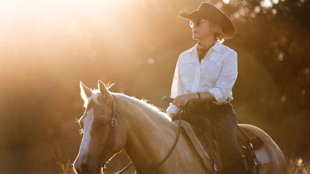 A woman in a white button-down shirt and black cowboy hat rides a pale palomino horse through golden late-afternoon light. Her expression is resolute as she gazes into the distance, sunlight flooding the frame from behind.