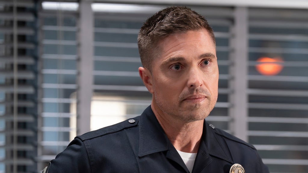 Close-up of a uniformed police officer with short hair and a serious expression, standing indoors in front of window blinds.