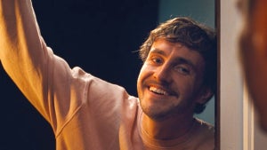 Paul Mescal in a still from "All of Us Strangers," smiling warmly while leaning out a doorway with one arm raised, wearing a peach sweatshirt in warm, low lighting.
